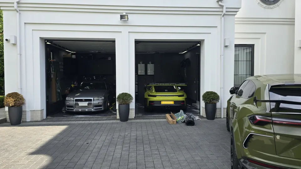 سيارة Nissan Skyline GT-R فضية اللون وسيارة بورش 911 GT3 صفراء متوقفة في المرآب، مع سيارة Lamborghini Urus باللون الأخضر الزيتوني متوقفة بالخارج.