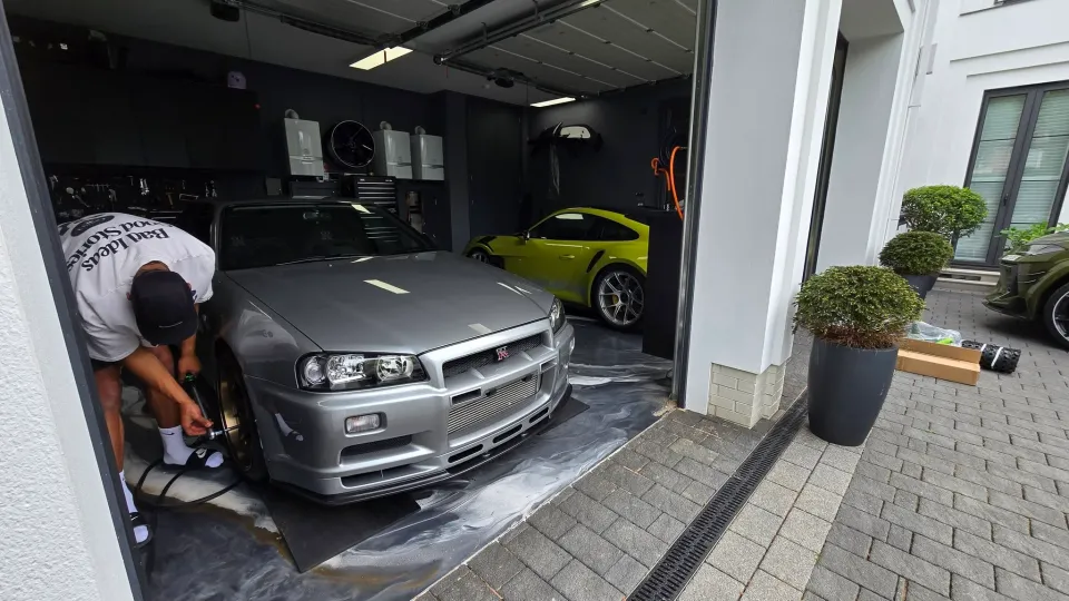 شخص يقوم بتنظيف سيارة Nissan Skyline R34 فضية داخل مرآب للسيارات مع سيارة بورش 911 GT3 RS صفراء في الخلفية.