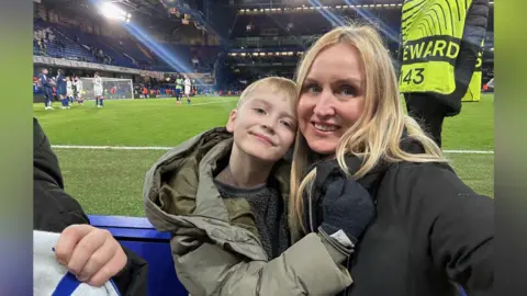 نشرة صبي ذو شعر أشقر، يرتدي سترة كاكي، يعانق امرأة ذات شعر أشقر وسترة سوداء. إنهم يشاهدون مباراة كرة قدم.