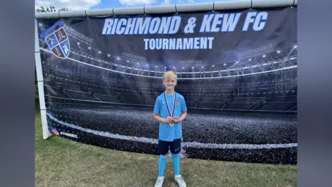 صبي ذو شعر أشقر، يرتدي قميص كرة قدم أزرق وسروالًا أسود وجوارب طويلة وأحذية رياضية بيضاء. وهو يقف أمام لافتة سوداء كبيرة. وهو يرتدي ميدالية حول رقبته.