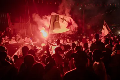 AFP عبر Getty Images أنصار السنغال يحملون شعلة أثناء احتفالهم في أوكام، داكار. الصورة مغمورة بالضوء الأحمر الناتج عن التوهج.